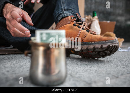 A picture that is focused on boots and a dirty hand of homeless man. He is reaching a cup that stands in front of him. There is a dollar in this metal Stock Photo