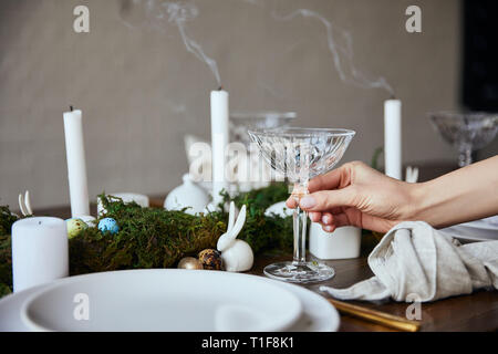 cropped view of woman putting crystal glass near plates, candles and moss on wooden table at home Stock Photo