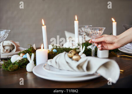 cropped view of woman putting crystal glass near plates, burning candles and moss on wooden table at home Stock Photo