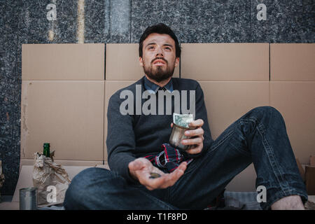 Adult beggar is sitting on cardboard and begging money. He is looking up. Man has some coins in one hand and a cup with some cash in another one. His Stock Photo