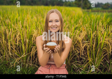 A woman is holding a cup of boiled rice in a wooden cup on the background of a ripe rice field Stock Photo