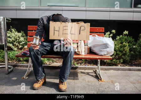 Guy is sitting on bench and holding a cardboard with help word on it. Also he has a cup with dollar bill in right hand. Homeless man is begging for Stock Photo