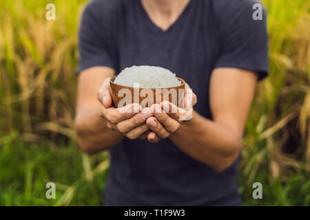 Man is holding a cup of boiled rice in a wooden cup on the background of a ripe rice field Stock Photo