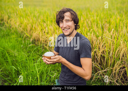 Man is holding a cup of boiled rice in a wooden cup on the background of a ripe rice field Stock Photo
