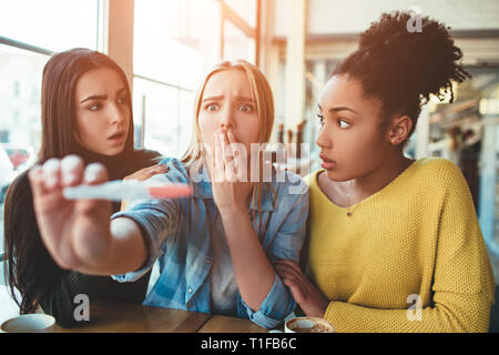 blonde girl is holding a pregnancy test that shows to her positive result of the test. She looks dissapointed, sad and trying not to cry. Her two best Stock Photo