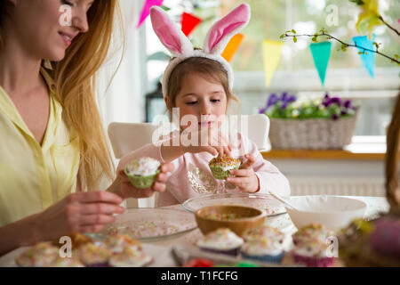 Mother and daughter celebrating Easter, cooking cupcakes, covering with glaze. Happy family holiday. Cute little girl in bunny ears. Stock Photo