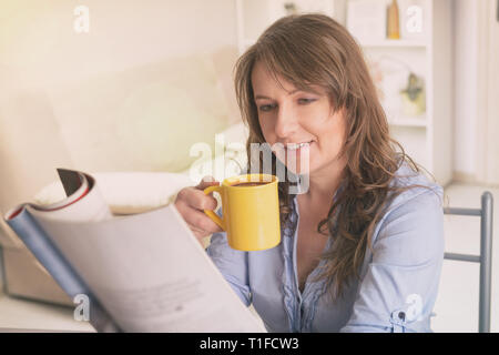 Beautiful young woman drinking morning coffee and reading newspaper at home Stock Photo