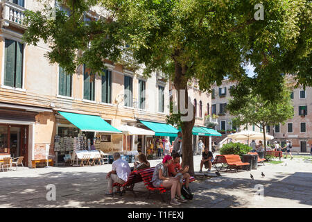 People sitting on benches in the shade of a green tree on a hot day in spring, Campo Santa Maria Nova, Cannaregion, Venice, Veneto, Italy Stock Photo