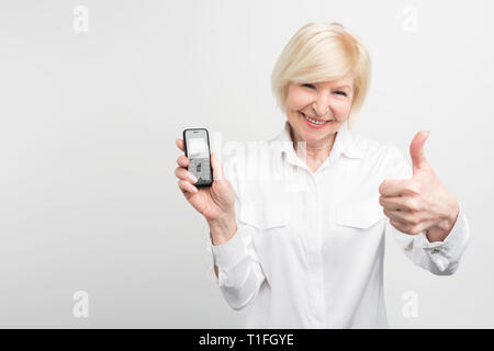 White-haired lady is holding an old phone. She prefers to use it instead of buying and using new one. This woman doesn't like new technologies Stock Photo