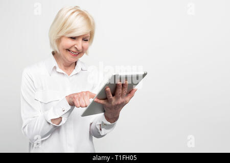 Old but happy woman is using a brand new tablet. She is testing it because she likes new technologies. Isolated on white background. Stock Photo