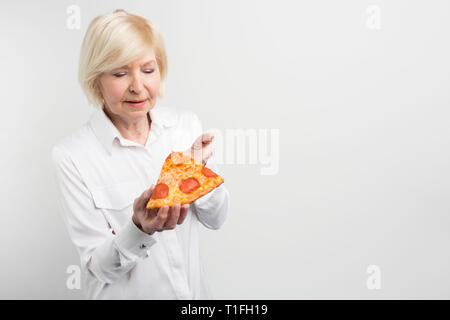 Old lady holding a big piece of pizza in her hands. She is trying to guess what ingredients are in it. Also she is thinking about eating that piece Stock Photo