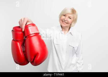 Joyful and nice old woman is holding boxing gloves in her right hand and smiling. She has what to do in her retirement. Isolated on white background. Stock Photo