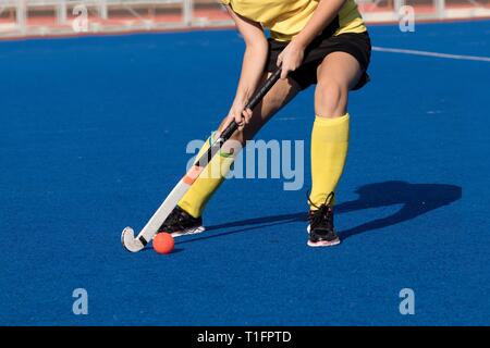 Young field hockey player in black and yellow uniform hitting the ball on blue water based turf during a game Stock Photo