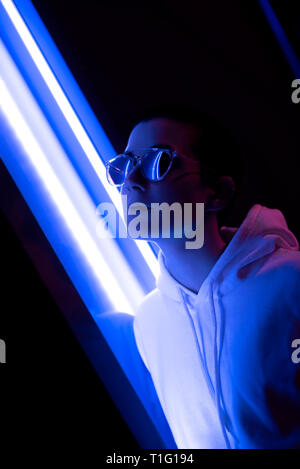 Colorful portrait of a young short haired woman wearing shiny sunglasses and standing in neon lights. Stock Photo