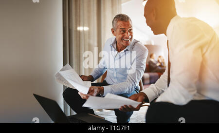 Two diverse businessmen smiling while sittingon chairs in a modern office discussing a project together Stock Photo