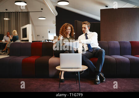 Two diverse businesspeople smiling while sitting together on a sofa in a modern office discussing paperwork Stock Photo