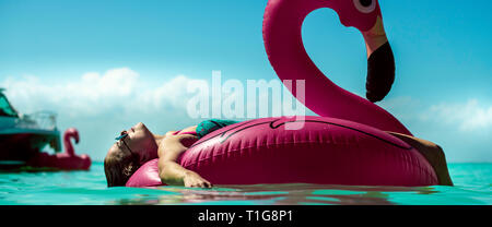Young woman resting for a tan in inflatable flamingo at a sunny day at sea with yatchs on the horizon. Stock Photo