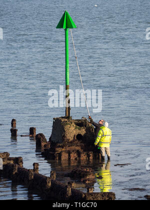 pair of two workmen men workers in high vis working maintaining painting repainting with extension pole brush roller a beach groyne marker navigation  Stock Photo