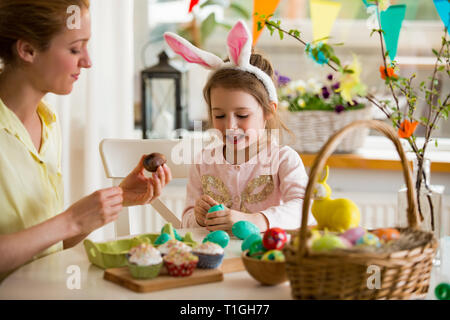 Mother and daughter celebrating Easter, eating chocolate eggs. Happy family holiday. Cute little girl with funny face in bunny ears laughing, smiling Stock Photo