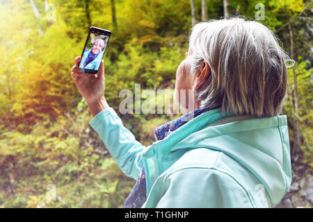 Nice attractive elderly mature woman with shiny grey hair taking photos and selfies outdoor in a Vintgar river canyon gorge on a beautiful sunny day Stock Photo