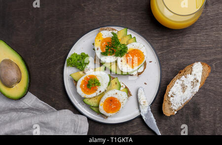 Two tasty toasts with avocado and boiled egg on white plate, grain bread with cream cheese and glass of orange juice near on wooden background Stock Photo