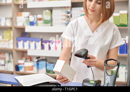 Pretty female pharmacist working in drugstore with medicaments. Woman holding medical box and scanning price of medicament using barcode scanner. Specialist standing at counter. Stock Photo