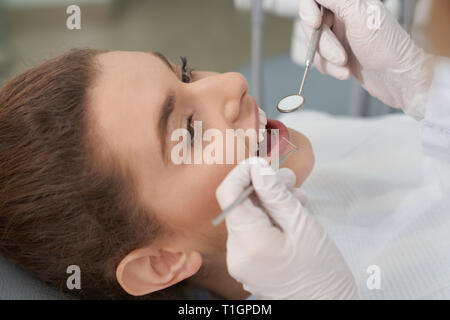 Closeup of pretty young woman lying on dental chair with open mouth and looking at doctor while she examining teeth. Client visiting dental office and treating tooth. Concept pof stomatology. Stock Photo