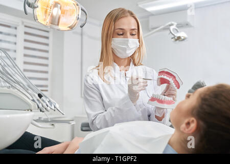 Dentist showing to patient dental teeth model. Client lying in dentist chair, consulting with doctor. Professional doctor wearing in white uniform, gloves and medical mask. Stock Photo