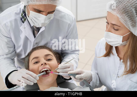 Beautiful woman with white teeth lying on dentist chair with opened mouth, looking at camera. Doctor holding restoration instruments. Stomatologist wearing in white medical masks and caps. Stock Photo