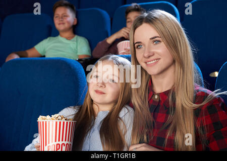 Beautiful woman sitting with little girl in cinema, enjoying premiere of film. Cheerful mother and daughter watching movie, girl holding popcorn bucket. Stock Photo