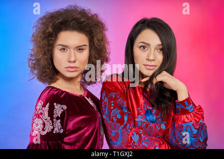 Models wearing in traditional red dresses posing. Charming girl with curly hair and elegant brunette looking at camera. Women wearing in embroidered clothes, atlas fabric. Stock Photo