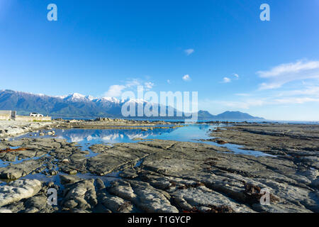 Snow-capped Kaikoura Mountains reflected in calm rockpool on mud-stone rock ledge Stock Photo