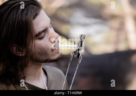 Portrait of a handsome young man with a beard, lighting a cigarette from charcoal, holding his by barbecue tongs. Stock Photo