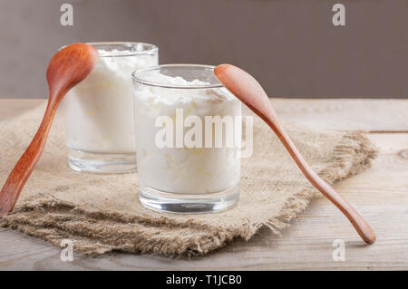 Two glasses with  homemade yogurt and spoons on burlap napkin on wooden table on beige background. Stock Photo