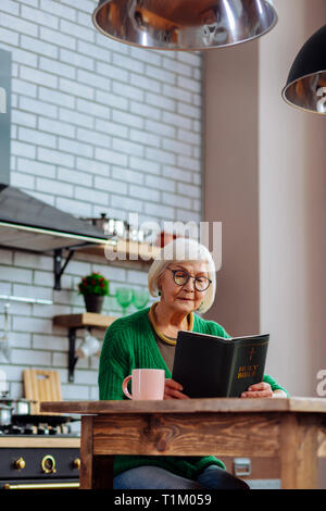 Elderly female busy with mindful reading of Bible at kitchen Stock Photo