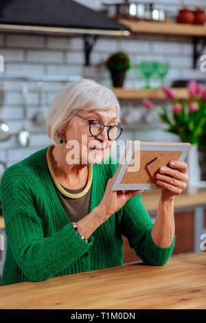 Aging lady lovingly looking at picture sitting at kitchen table Stock Photo