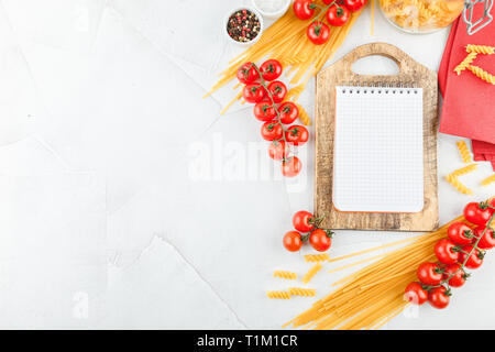 Notebook on wooden cutting board with different types of dry pasta and tomatoes on white wooden background, top view. Copy space. Flat lay. Stock Photo