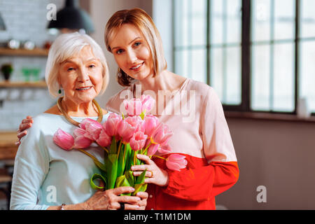 Beautiful mature daughter giving tulips to wrinkled grey-haired mother Stock Photo