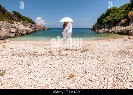 Romantic view of couple in white clothes cuddle under white umbrella, standing on rocky beach and looking on picturesque landscape of blue sea. Concep Stock Photo