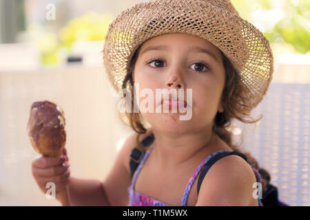 Little girl with straw hat and a sundress enjoys the summer heat eating a refreshing cone of chocolate ice cream, it melts on her hand and she has the Stock Photo