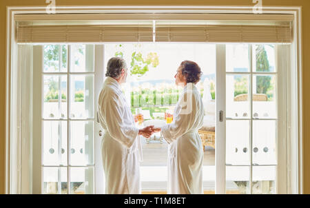 Mature couple in spa bathrobes drinking mimosas at hotel patio door Stock Photo