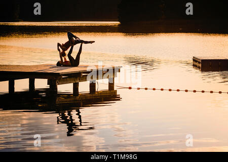 Women practicing acro yoga on lakeside dock at dusk Stock Photo