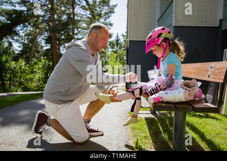Father helping his daughter in helmet to wear protective pads for roller skates. Happy family spending time together. Sunny summer day on suburb Stock Photo