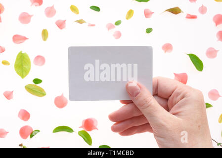 Woman holding blank white business card mock up in hand over white background with blooming springtime flower petals and leaves scattered around Stock Photo