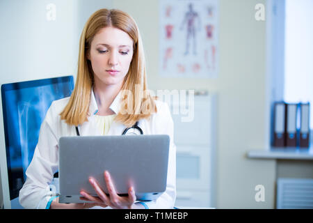 Portrait of friendly confident female doctor in hospital working on laptop. Enthusiastic medical staff at work. Female doctor in diagnostics room. Stock Photo