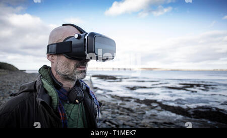 A front-view shot of a mature caucasian man wearing a virtual reality headset while standing on a beach in the United Kingdom, he is having fun. Stock Photo