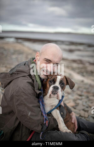 A front-view shot of a mature caucasian man sitting on a beach with his brown and white boxer dog, they are embracing eachother on the cold day in the Stock Photo