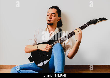 Young millenial man sitting on the floor of home playing an electric guitar. Stock Photo