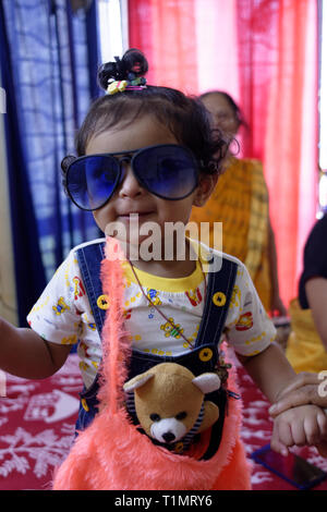 A beautiful Indian  infant girl child posing in a funny mood wearing a blue big sun glass in a homely environment. Stock Photo