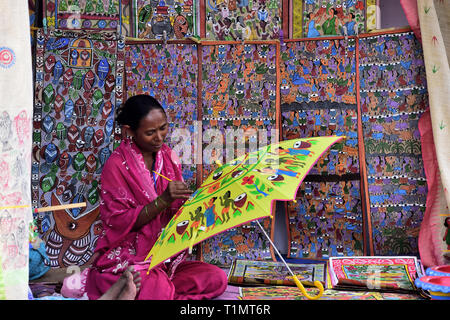 Indian rural woman artist is painting an umbrella sitting by wall with hanging decorations Stock Photo